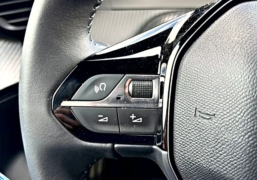 Gros plan sur les commandes gauche du volant en cuir noir du Peugeot 2008 gris Artense, avec boutons de volume et appel mains libres.