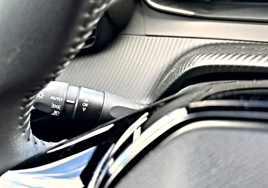 Gros plan sur le volant noir et la commande des phares du Peugeot 2008 1.2 100 Allure gris Artense, intérieur moderne.
