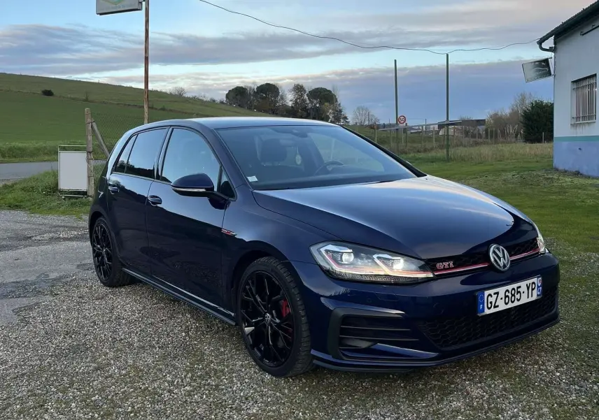 Volkswagen Golf VII GTI Performance bleu lapiz métal vue 3/4 avant droit sur parking gravier.