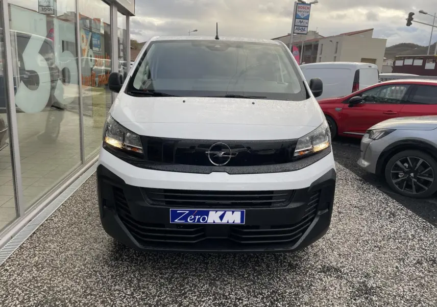 Vue frontale d'un Opel Vivaro blanc avec calandre noire et phares allumés sur parking extérieur.