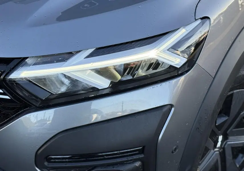 Gros plan sur le phare avant droit allumé du Dacia Jogger Gris Schiste avec détails de la calandre et de la roue.