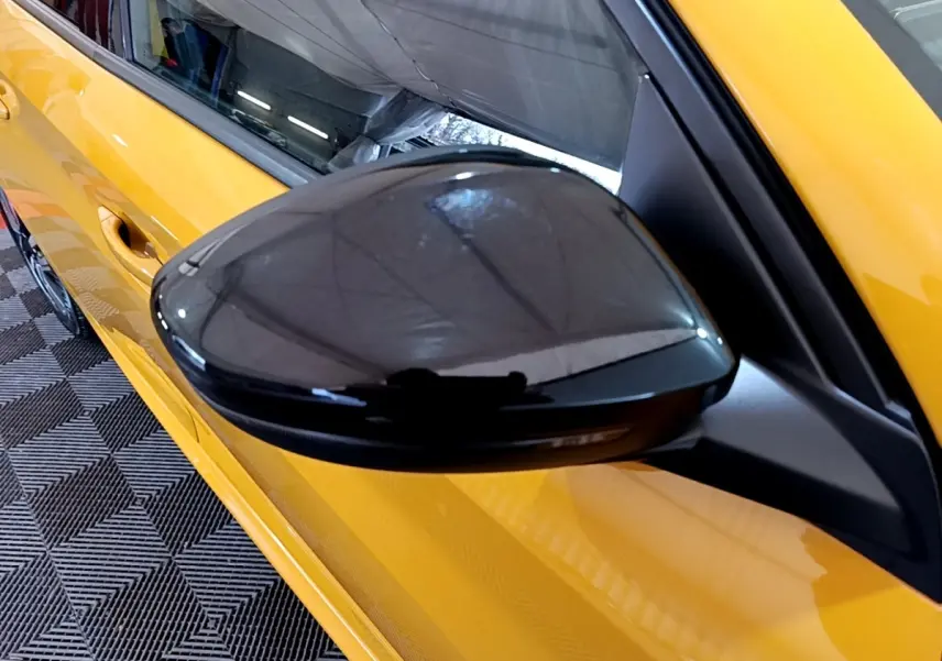 Rétroviseur noir côté gauche d’une Peugeot 208 jaune vue en léger angle depuis l’avant.