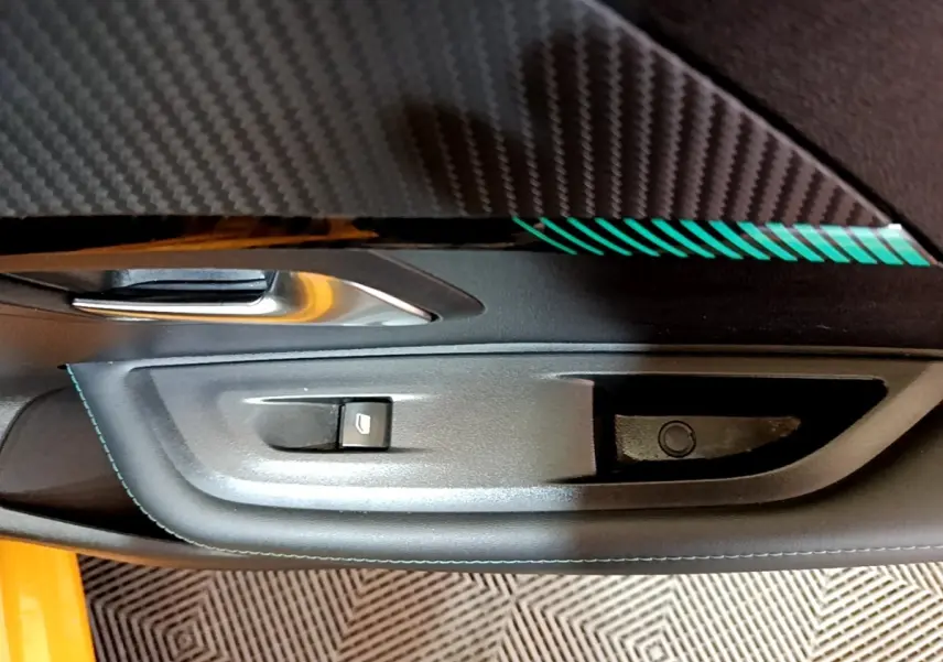 Gros plan sur la poignée intérieure côté gauche d’une Peugeot 208 jaune avec surpiqûres bleues et insert carbone.