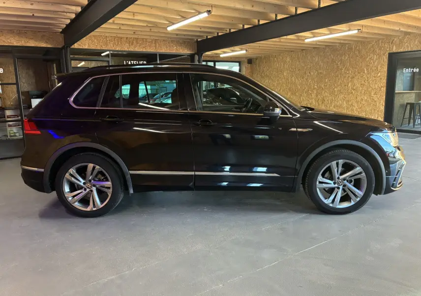 Volkswagen Tiguan noir vu de profil côté gauche, avec jantes alu et barres de toit dans un garage.
