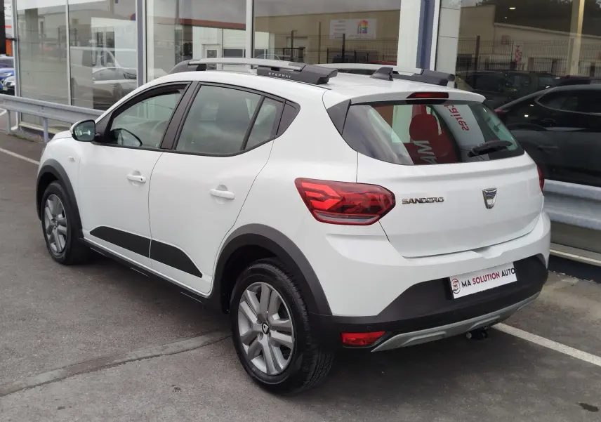 Vue 3/4 arrière droite d'une Dacia Sandero Stepway blanche Glacier avec barres de toit et protections noires.