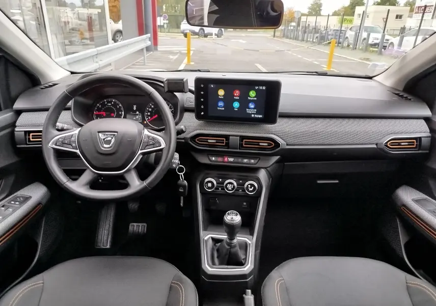 Vue intérieure avant de la Dacia Sandero Stepway 2021, tableau de bord noir avec touches orange et écran tactile central.
