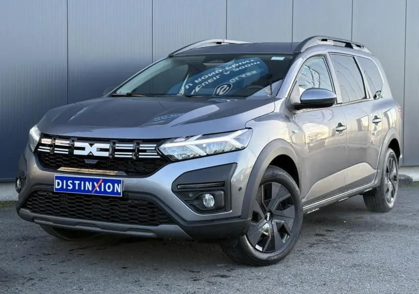 Dacia Jogger Gris Schiste en 3/4 avant droit, avec calandre noire, jantes noires et barres de toit longitudinales.