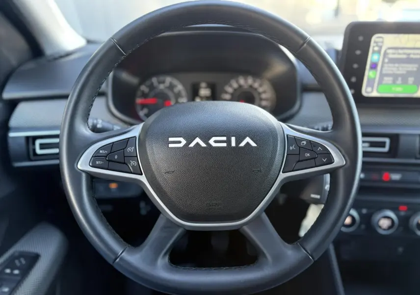 Gros plan sur le volant noir Dacia avec commandes intégrées, tableau de bord flou et écran tactile visible à droite.
