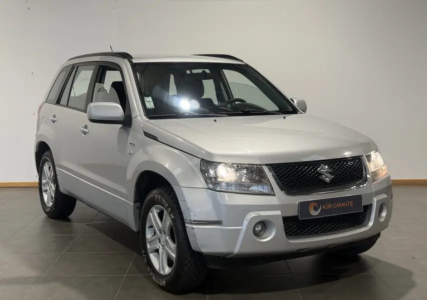 Suzuki Grand Vitara gris 2007 en 3/4 avant droit avec calandre noire et phares allumés en intérieur showroom.