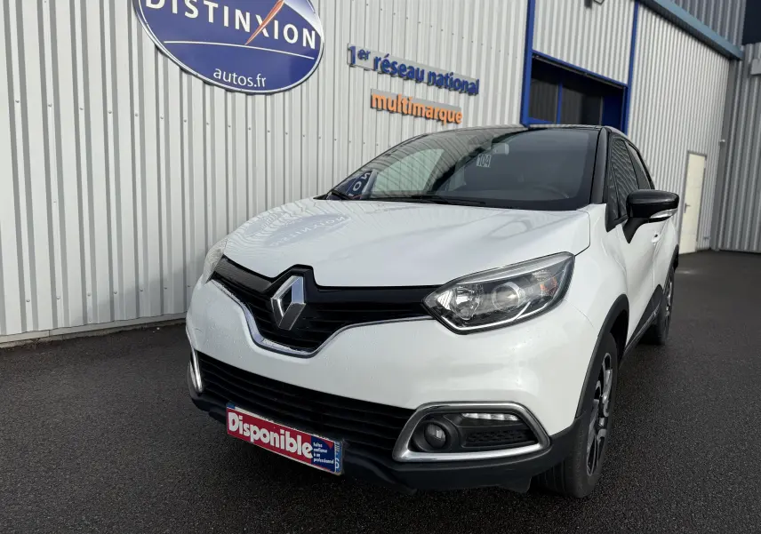 Renault Captur blanc en 3/4 avant droit, avec calandre noire et feux LED, stationné devant un bâtiment industriel.