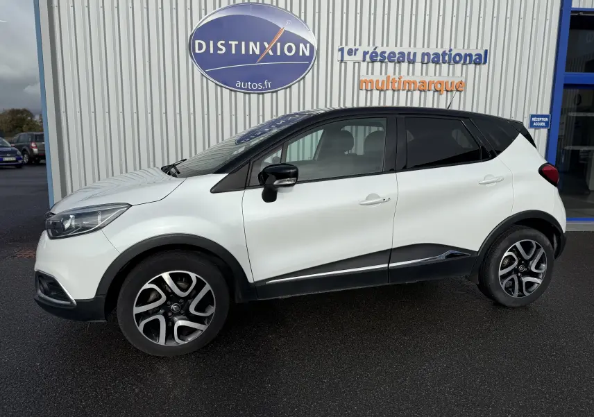 Profil côté gauche d'un Renault Captur blanc 2017 avec toit noir et jantes alliage bicolores devant un bâtiment Distinxion.