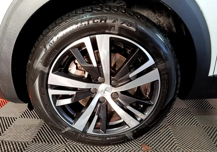 Gros plan sur la roue avant droite d’un Peugeot 3008 blanc, mettant en valeur la jante alliage bicolore et le logo Peugeot central.