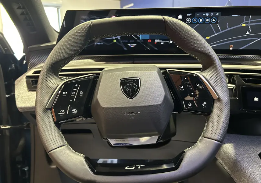 Vue rapprochée du volant multifonction cuir perforé du Peugeot 3008 III HYBRID GT avec tableau de bord numérique en arrière-plan.