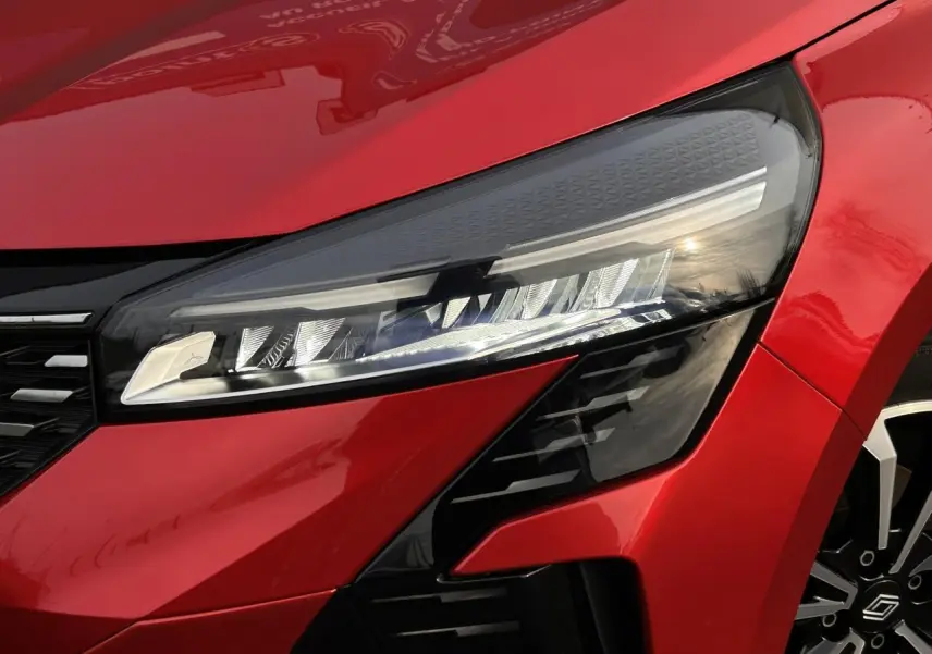 Gros plan sur le phare avant droit et la calandre d'une Renault Clio 2025 Rouge Désir avec jante alliage visible.