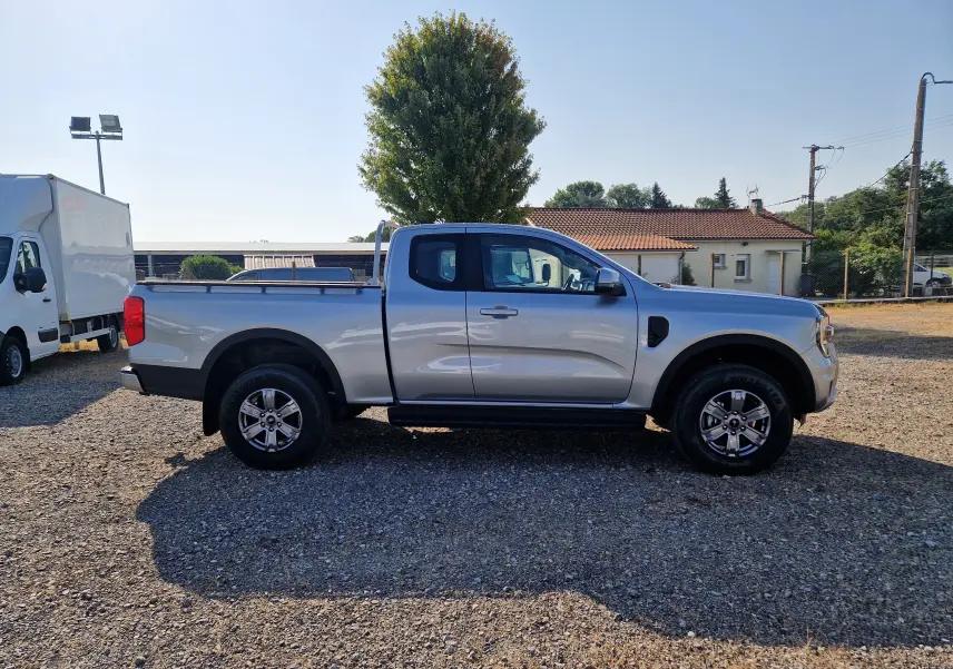 Ford Ranger Super Cabine gris iconic vu de profil côté gauche sur un terrain gravillonné avec maison en arrière-plan.