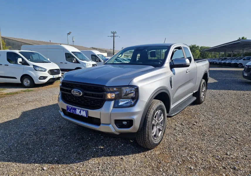Ford Ranger Super Cabine gris iconic vu en 3/4 avant droit sur un terrain gravillonné avec plusieurs véhicules utilitaires blancs en arrière-plan.