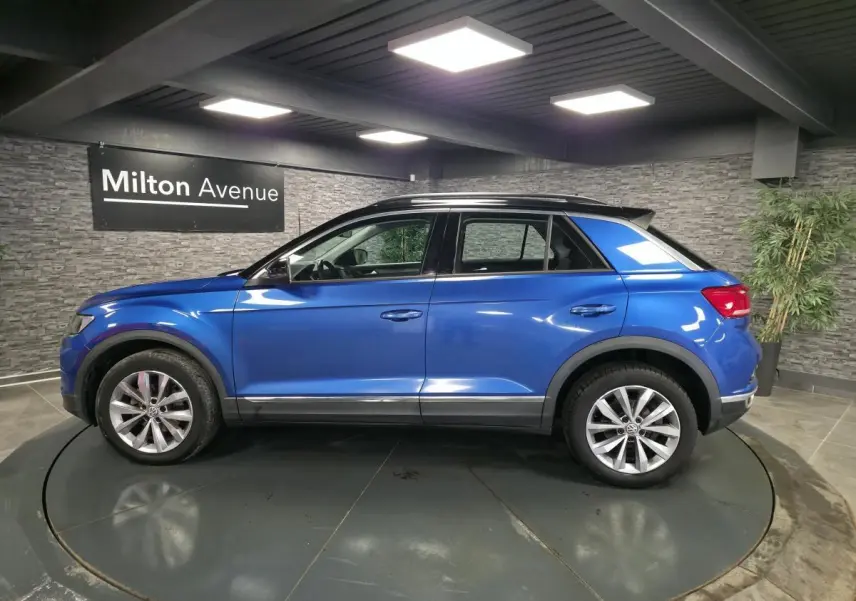 Vue de profil côté gauche d'un Volkswagen T-Roc bleu foncé avec jantes alliage et toit noir en intérieur showroom.