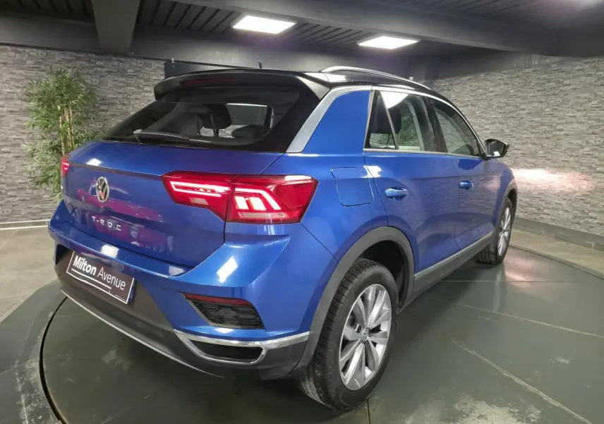 Vue 3/4 arrière droite d'un Volkswagen T-Roc bleu foncé avec toit noir et jantes alliage argentées en intérieur showroom.