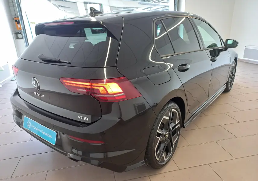 Vue 3/4 arrière droite d'une Volkswagen Golf 1.5 eTSI 150 DSG7 R-Line Edition noire Ébène avec feux allumés en intérieur showroom.