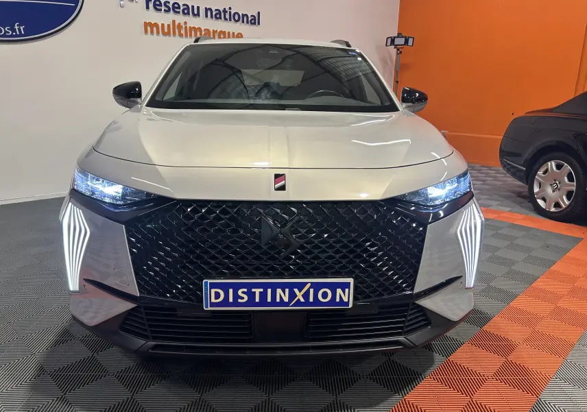 Vue frontale d’un DS7 2024 couleur Cristal Pearl avec calandre noire et feux LED distinctifs allumés en intérieur showroom.