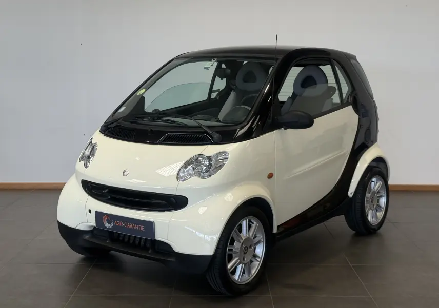 Vue 3/4 avant d'une Smart Fortwo Coupé beige et noire de 2004 avec toit panoramique en intérieur showroom.