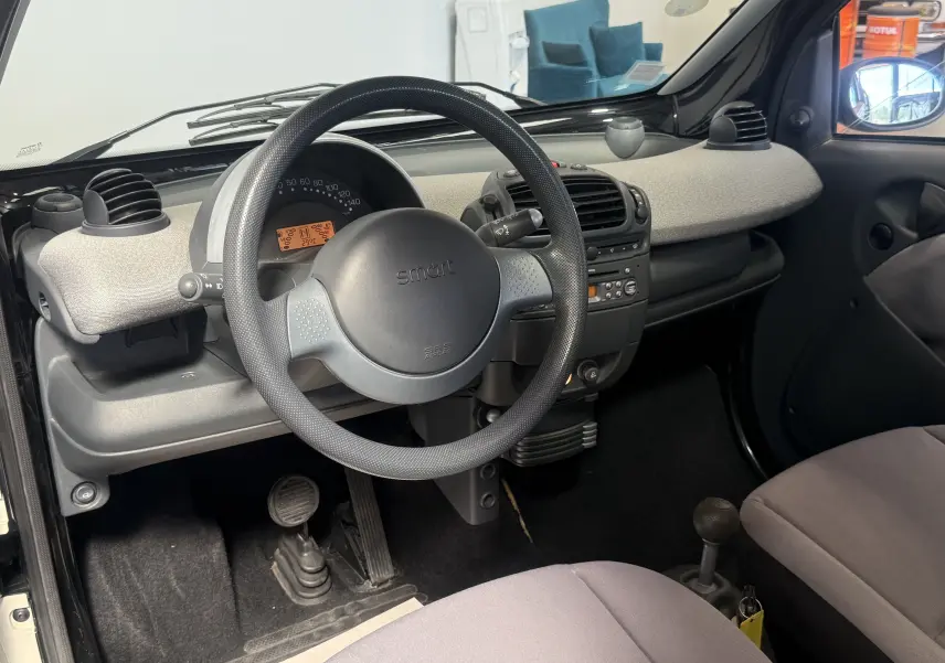 Vue intérieure du poste de conduite de la Smart Fortwo beige 2004, avec volant noir et tableau de bord gris clair.