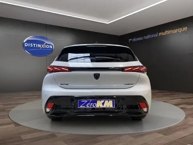 Vue arrière d'une Peugeot 308 Hybrid blanche 2026, avec feux LED fins et logo noir central sur fond showroom.