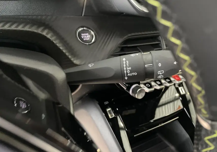 Détail intérieur du Peugeot 208 2023 montrant la commande essuie-glace, bouton start/stop et partie du volant avec surpiqûres vertes.