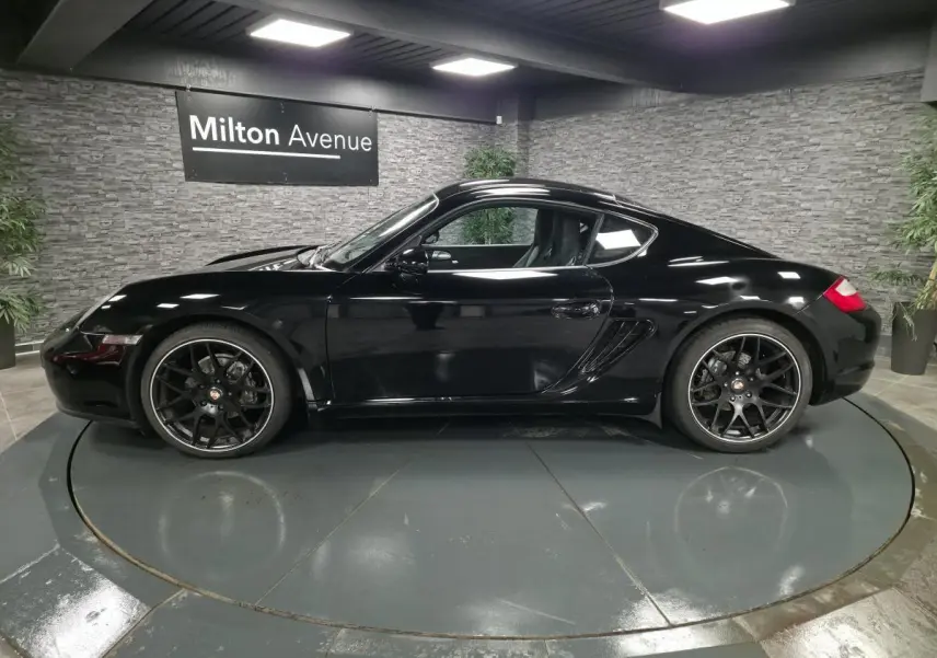 Profil droit d'une Porsche Cayman 2.7i noire de 2008 avec jantes SportDesign noires de 19 pouces, en intérieur showroom.