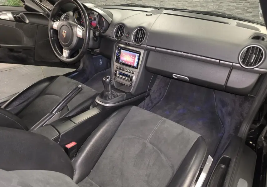 Vue intérieure côté passager d’une Porsche Cayman 2.7i noire de 2008 avec tableau de bord noir et levier de vitesses manuel.