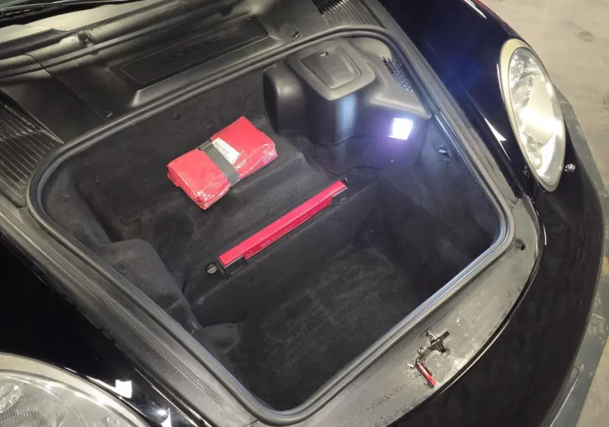 Vue plongeante sur le coffre avant noir d'une Porsche Cayman 2.7i 2008 avec trousse de secours rouge visible.