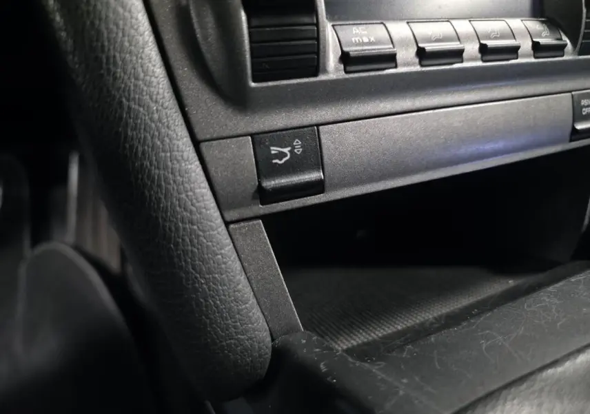Gros plan sur la console centrale noire de la Porsche Cayman 2.7i 2008, montrant les boutons de commande et un rangement ouvert.