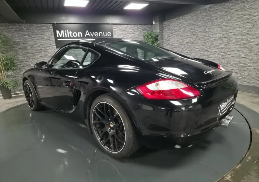 Vue 3/4 arrière droite d'une Porsche Cayman 2.7i noire avec jantes SportDesign noires de 19 pouces et double sortie d'échappement centrale.