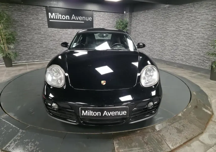 Vue avant d'une Porsche Cayman 2.7i noire, avec phares bi-xénon et plaque Milton Avenue visible.
