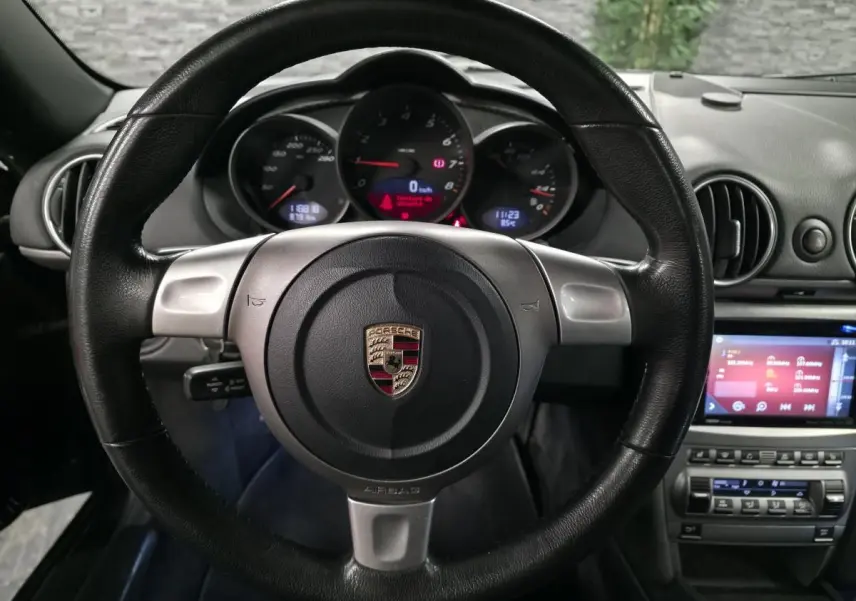 Vue intérieure centrée sur le volant noir en cuir et le tableau de bord avec compteurs ronds du Porsche Cayman 2.7i 2008.
