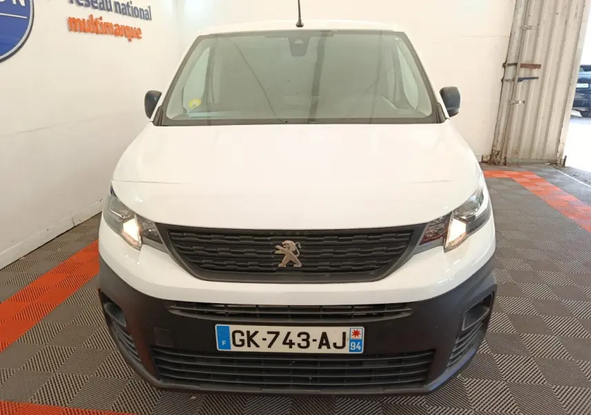 Vue frontale d’un Peugeot Partner blanc 2022 avec calandre noire et phares allumés en intérieur showroom.
