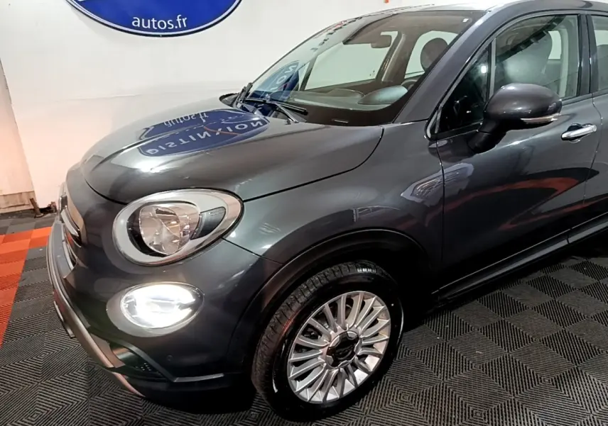 Vue 3/4 avant droit d'une FIAT 500X City Cross gris foncé avec phares et feux diurnes allumés en intérieur showroom.