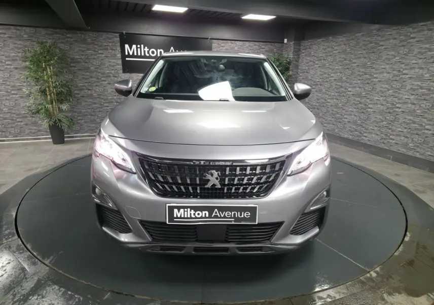 Vue avant frontale d'un Peugeot 3008 gris métallisé avec calandre noire et phares allumés en intérieur showroom.