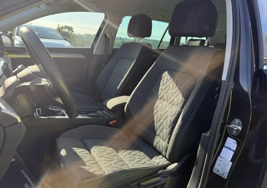 Vue intérieure côté conducteur de la Volkswagen Passat SW 2023, sièges tissu noir avec surpiqûres blanches et accoudoir central.