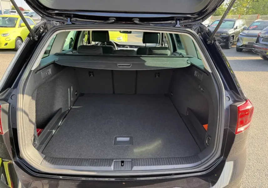 Coffre ouvert vu de l'arrière d'une Volkswagen Passat SW noire intense nacrée, espace de chargement vide et tapis gris.
