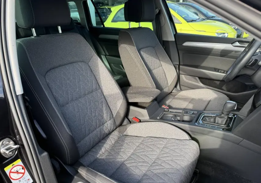 Vue intérieure côté conducteur de la Volkswagen Passat SW 2023, sièges tissu gris avec accoudoir central noir.