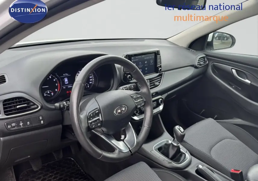 Vue intérieure côté conducteur de la Hyundai i30 2023, tableau de bord moderne avec écran tactile et levier de vitesses manuel.