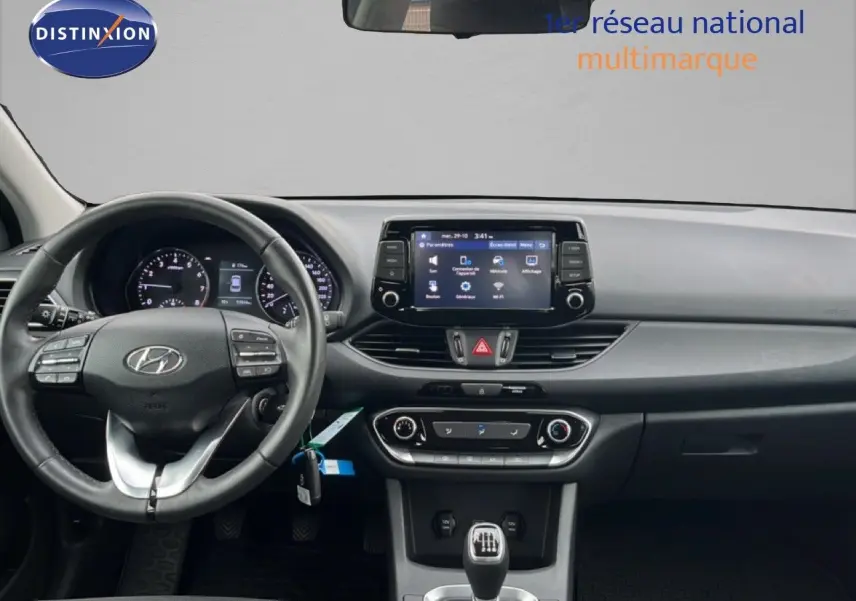 Vue intérieure frontale du tableau de bord et volant de la Hyundai i30 blanche nacrée, avec écran tactile et boîte manuelle.