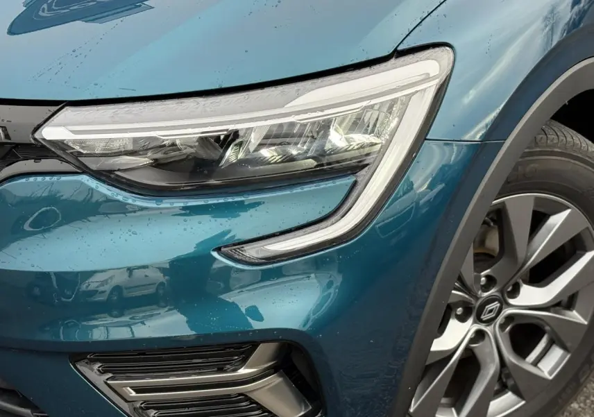 Gros plan sur le phare avant droit et la roue alliage grise du Renault Arkana Bleu Zanzibar sous la pluie.