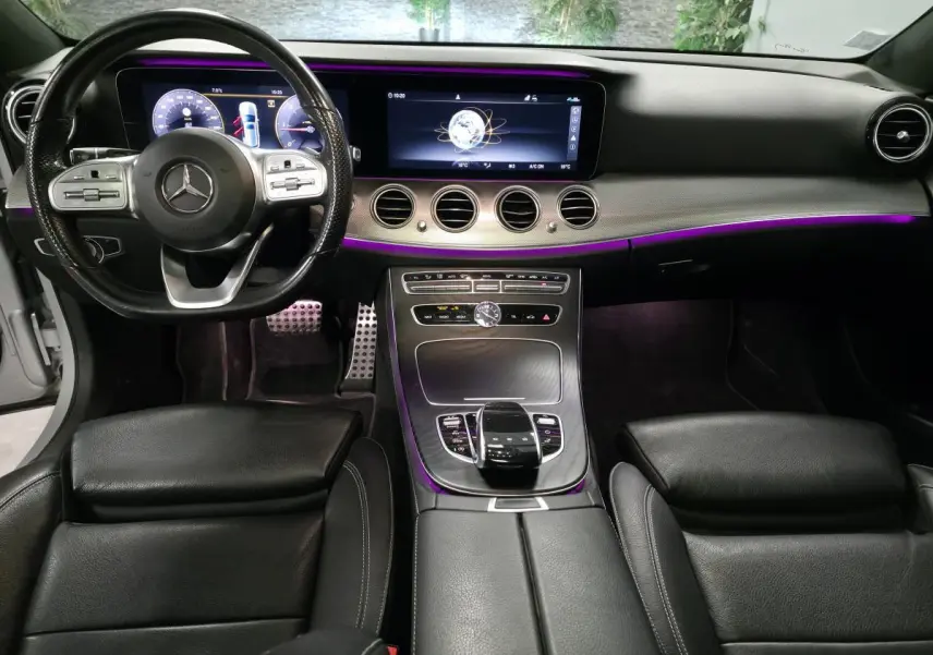 Vue intérieure avant de la Mercedes Classe E Break 2020, tableau de bord noir avec éclairage d'ambiance violet et volant multifonction.