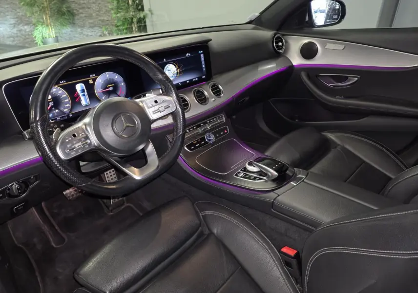 Vue intérieure côté conducteur de la Mercedes Classe E Break 2020, avec éclairage d’ambiance violet et finitions bois brun.