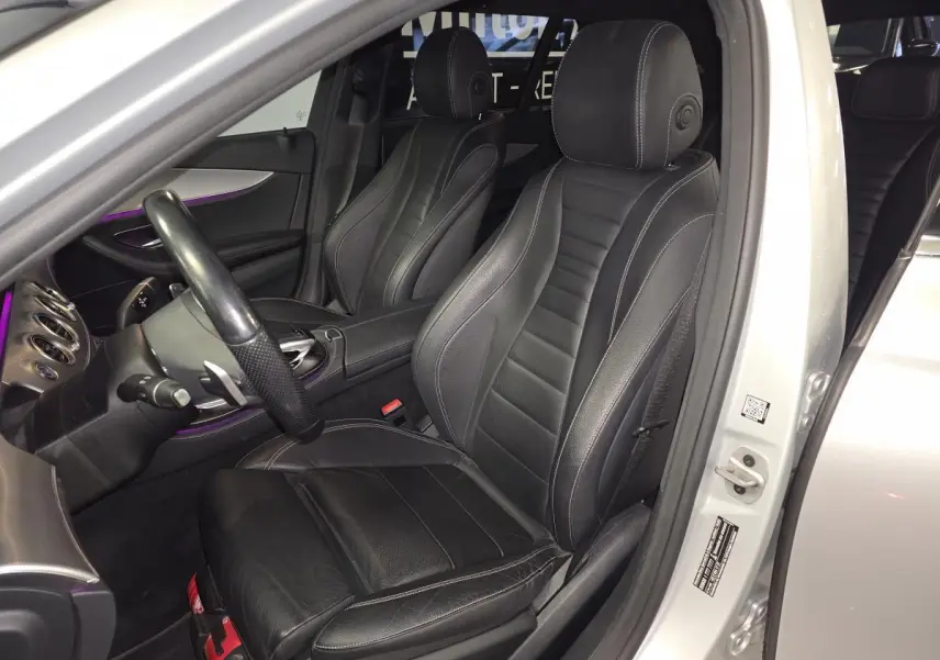 Vue intérieure côté conducteur d'une Mercedes Classe E Break gris, sièges cuir noir avec surpiqûres blanches et éclairage d'ambiance violet.