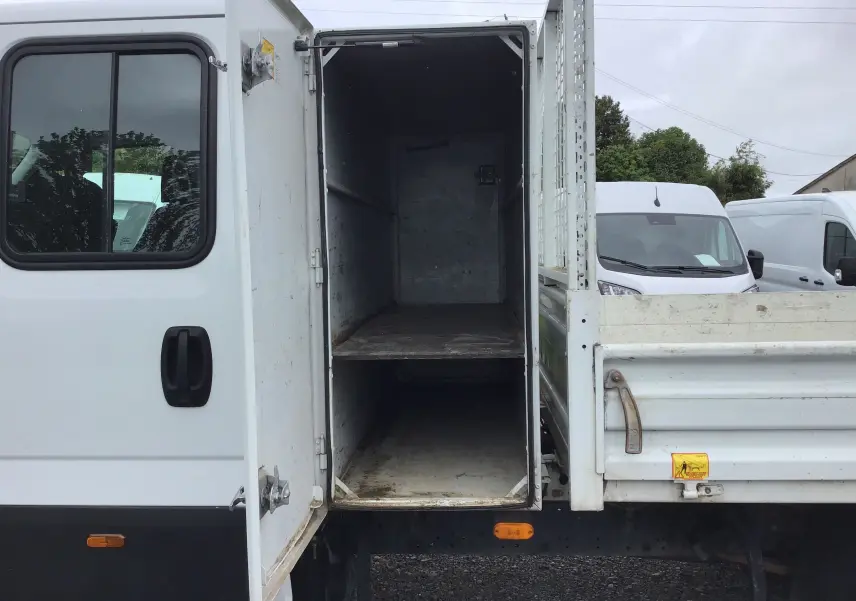 Vue latérale gauche d'un Iveco Daily blanc avec benne et coffre ouvert montrant l'intérieur vide du rangement.