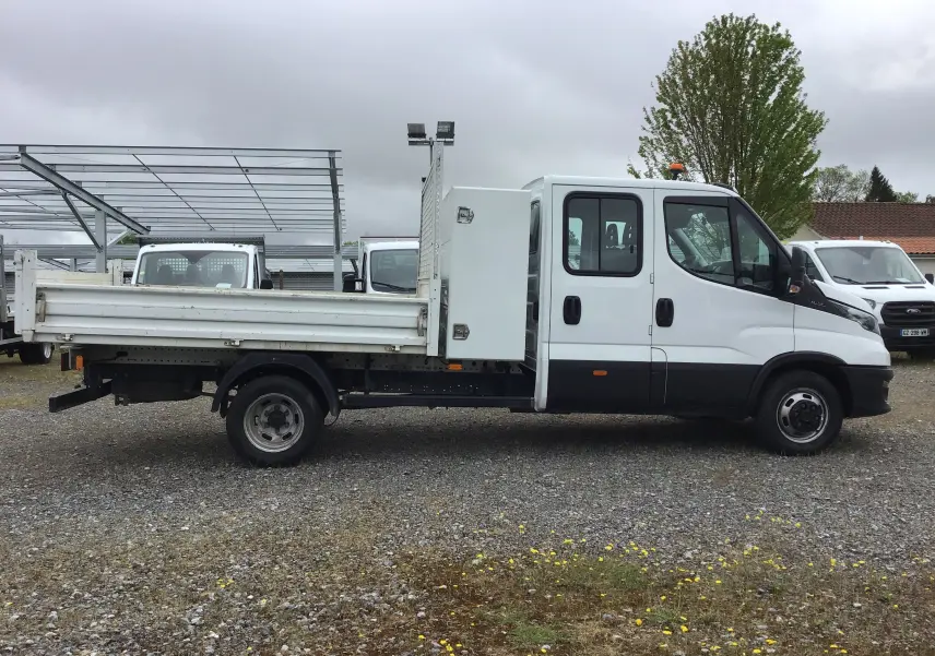 Vue de profil côté gauche d'un Iveco Daily blanc avec benne, coffre latéral et cabine double sur un sol gravillonné.