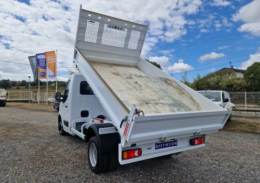 Vue 3/4 arrière droite d’un Renault Master blanc avec benne relevée et roues jumelées sur un sol gravillonné.